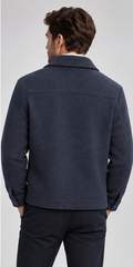 A man viewed from the back wearing a dark navy textured jacket and dark trousers against a light gray studio background.