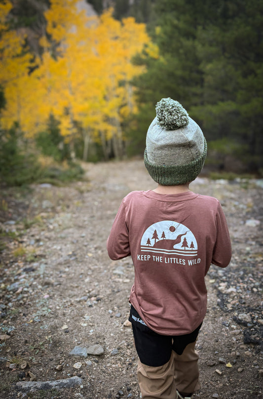 Person wearing a brown shirt with a logo and text, standing on a trail with autumn trees in the background.