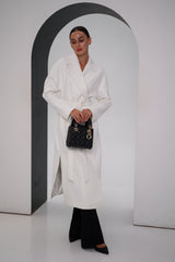 Woman wearing a white coat and black pants, holding a black handbag, against a plain background.