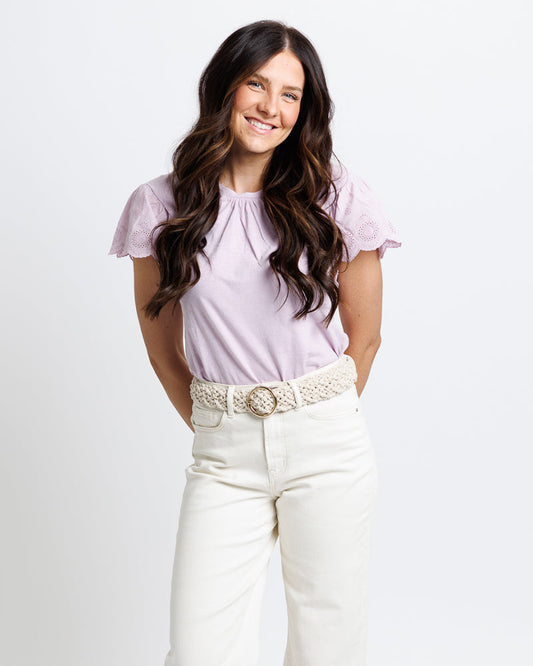 Woman wearing a light purple top and white pants with a belt on a plain background