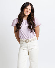 Woman wearing a light purple top and white pants with a belt on a plain background