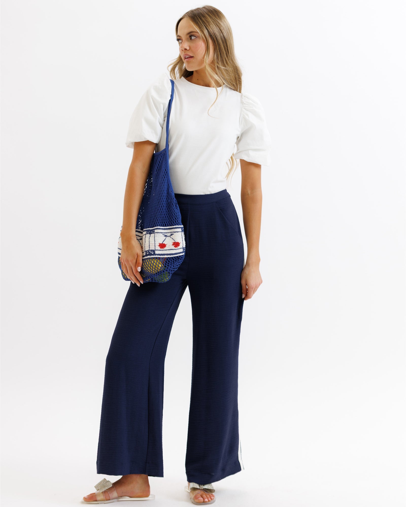 Woman wearing a white top and navy pants holding a blue bag on a white background