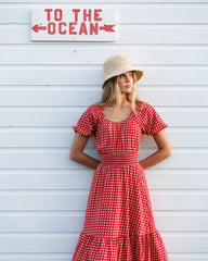 Woman in a red checkered dress and hat standing against a white wooden wall with 'To the Ocean' sign.