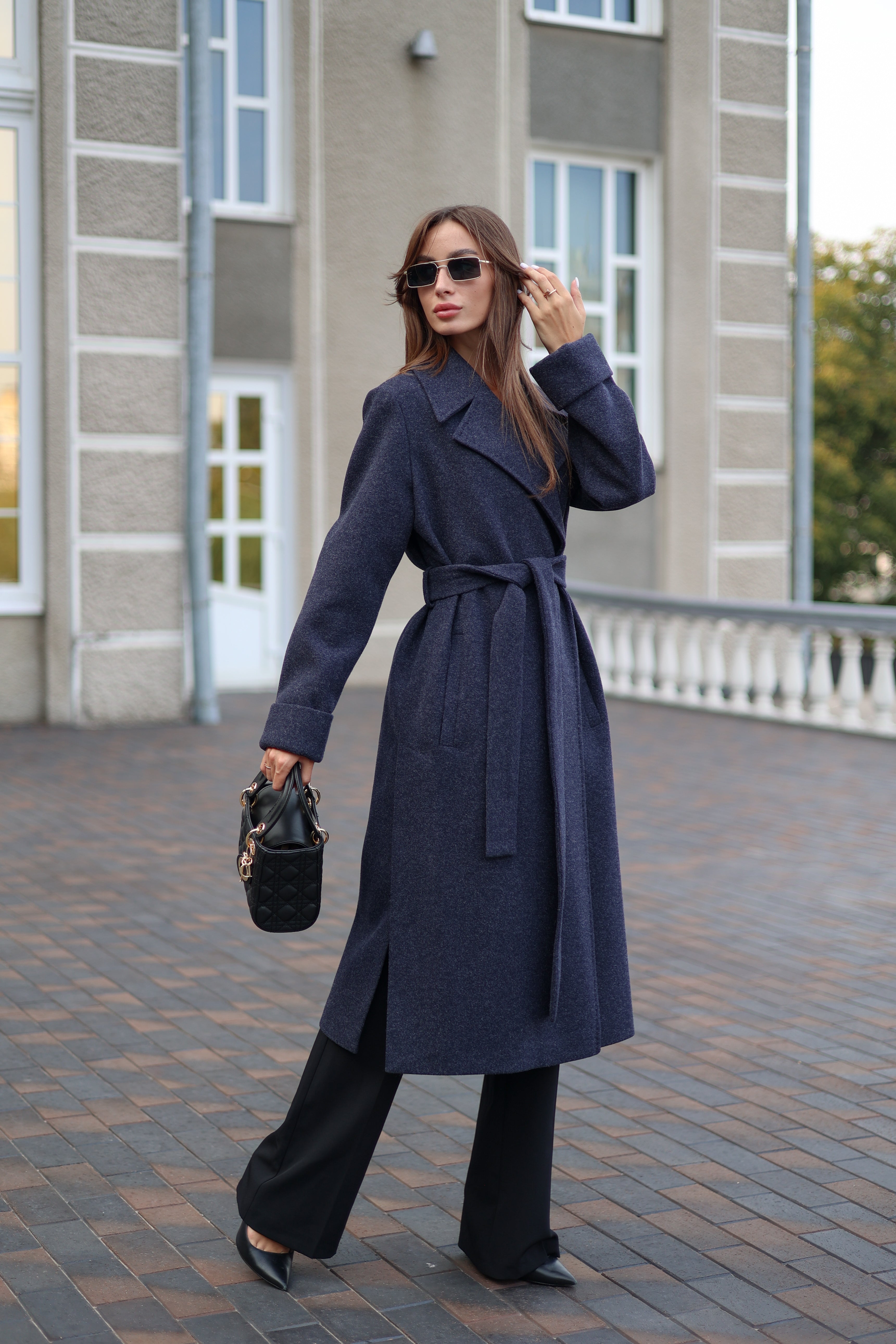 Woman wearing a long navy coat with a belt, holding a black handbag, standing on a paved walkway.