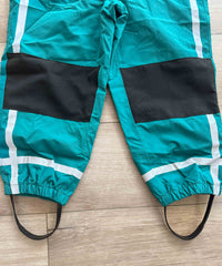 Teal rain pants with black patches and white reflective stripes on a wooden floor.