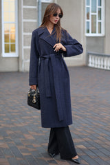 Woman wearing a long navy coat with a belt, holding a black handbag, standing on a paved walkway.