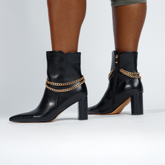 Black ankle boots with gold chain details worn by a person on a white background
