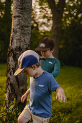 Two children standing outdoors near a tree, wearing caps and casual clothing.