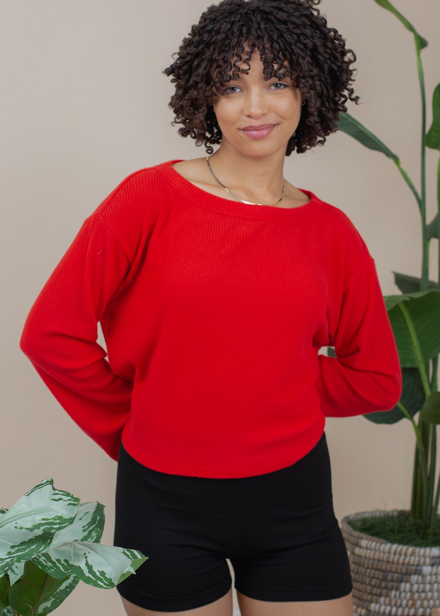 Woman wearing a red sweater and black shorts standing in front of plants.