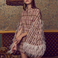 A woman wearing a brown checkered wool coat with feather-like trim on the sleeves, standing in a room with ornate wallpaper.