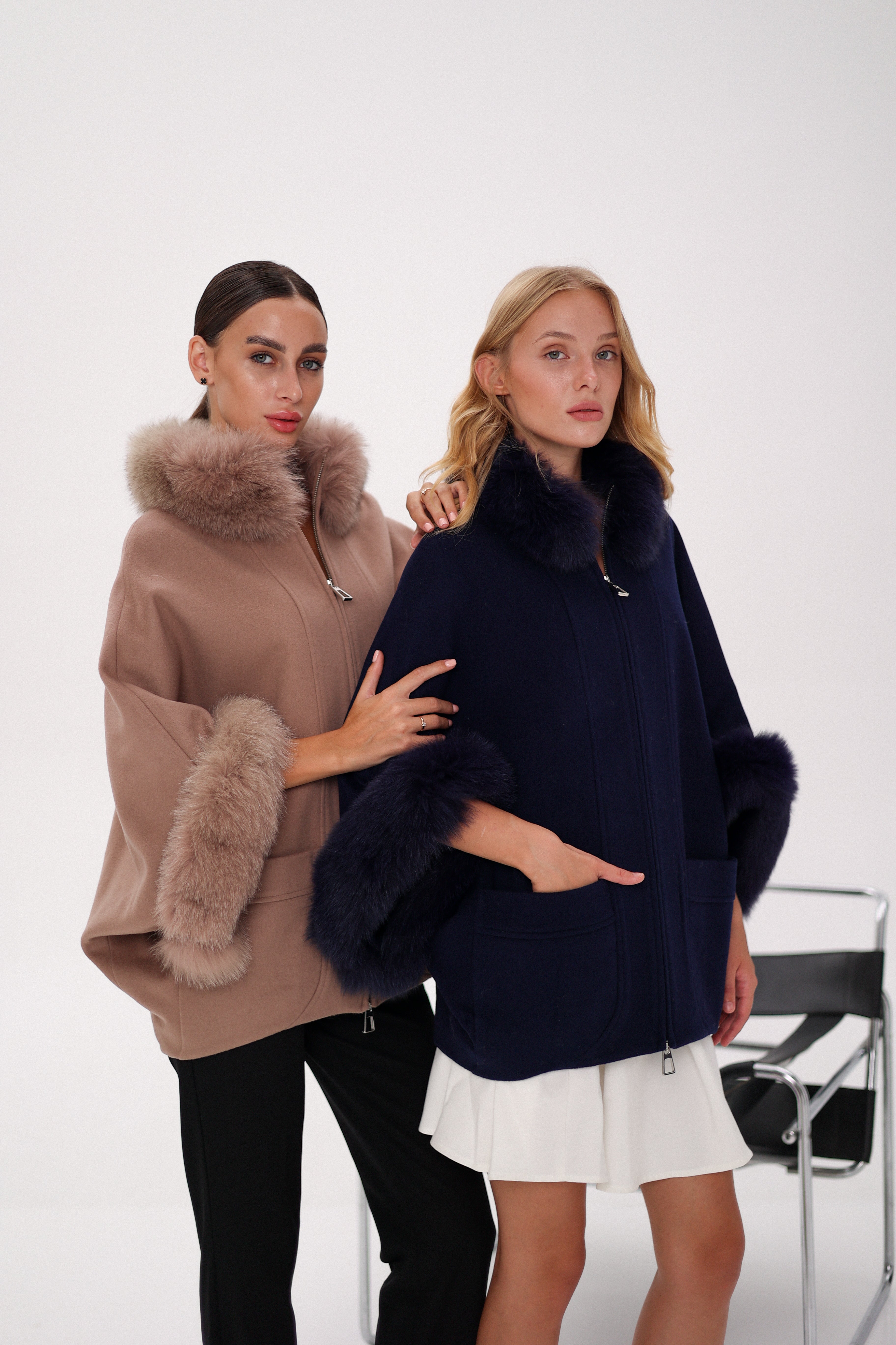Two women wearing fur-trimmed ponchos against a white background