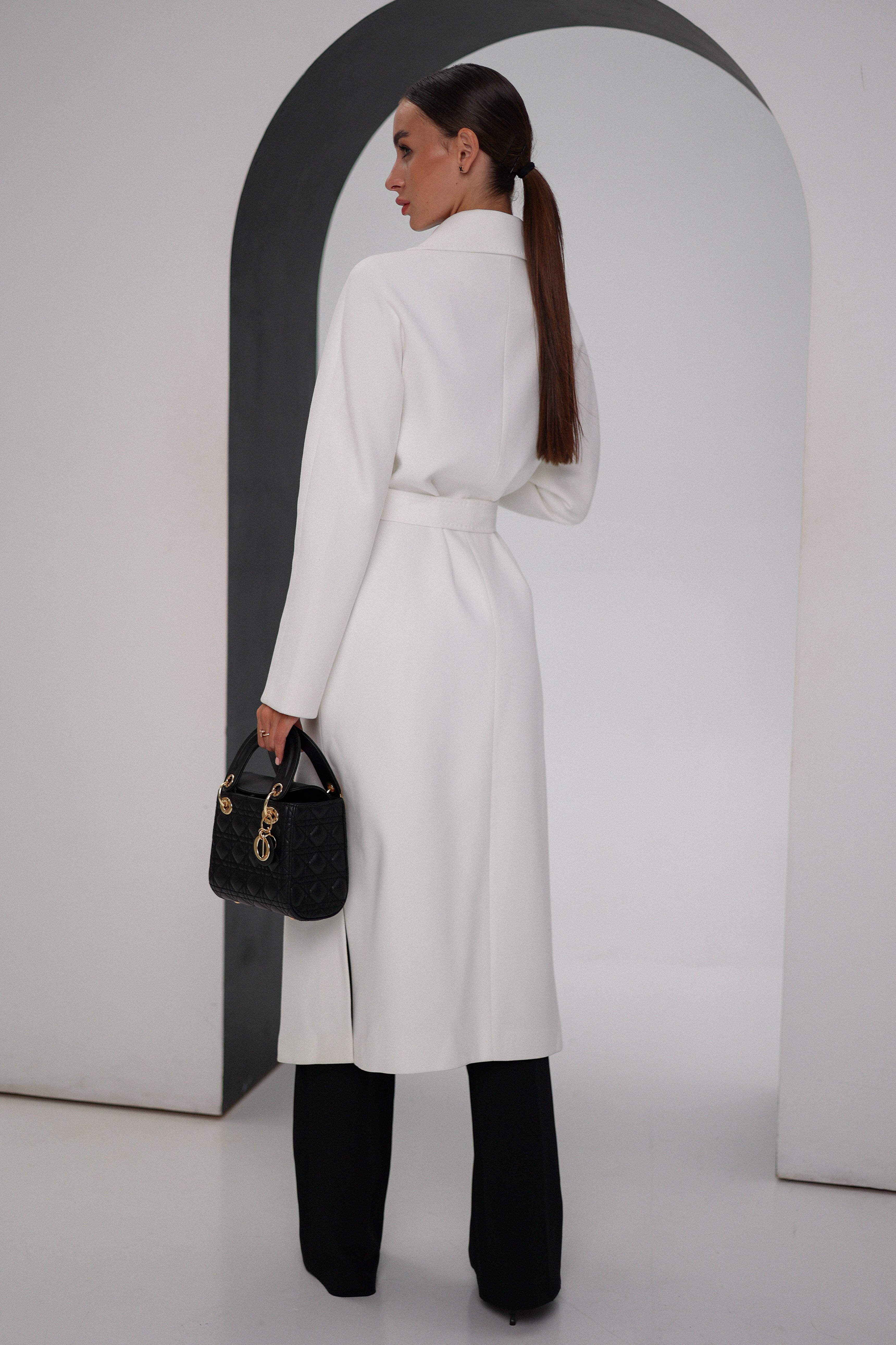 Woman wearing a white coat and black pants, holding a black handbag, against a plain background.