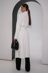 Woman wearing a white coat and black pants, holding a black handbag, against a plain background.