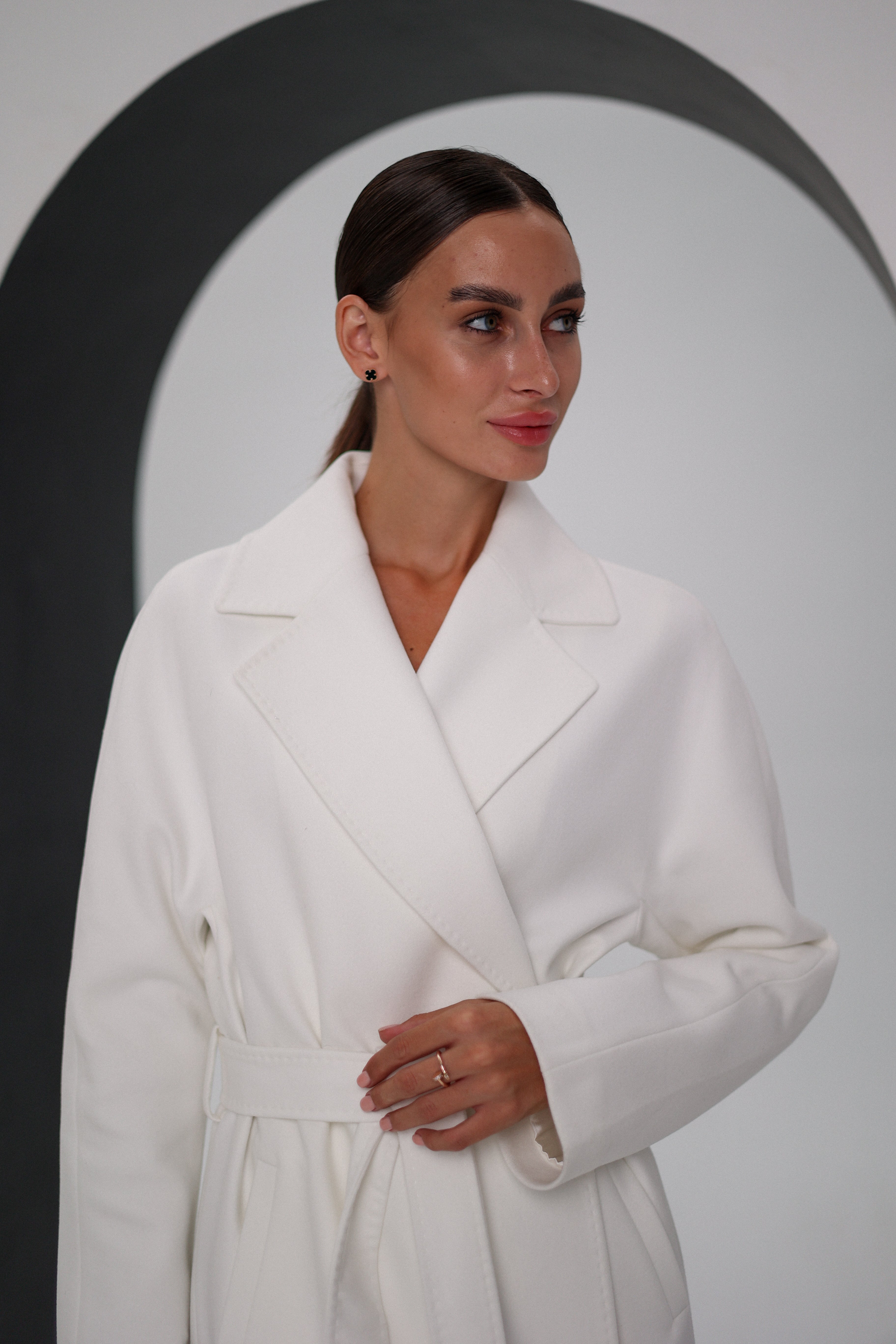 Woman wearing a white coat and black pants, holding a black handbag, against a plain background.