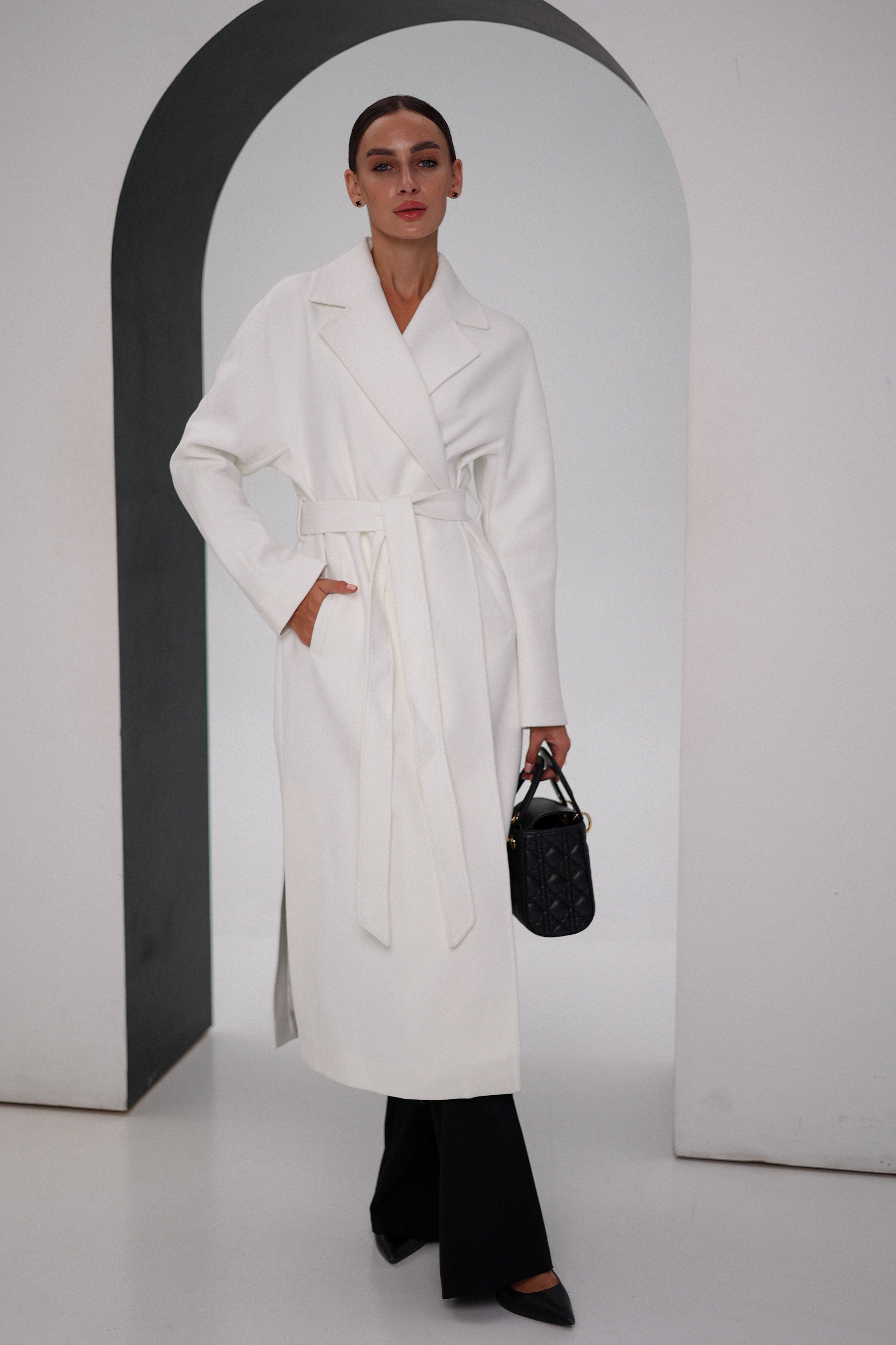 Woman wearing a white coat and black pants, holding a black handbag, against a plain background.