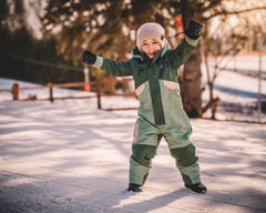 Child in a green snowsuit standing on a snowy path with trees in the background