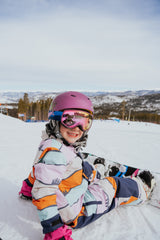 Person in colorful winter gear sitting on a snowy slope with a helmet and goggles.