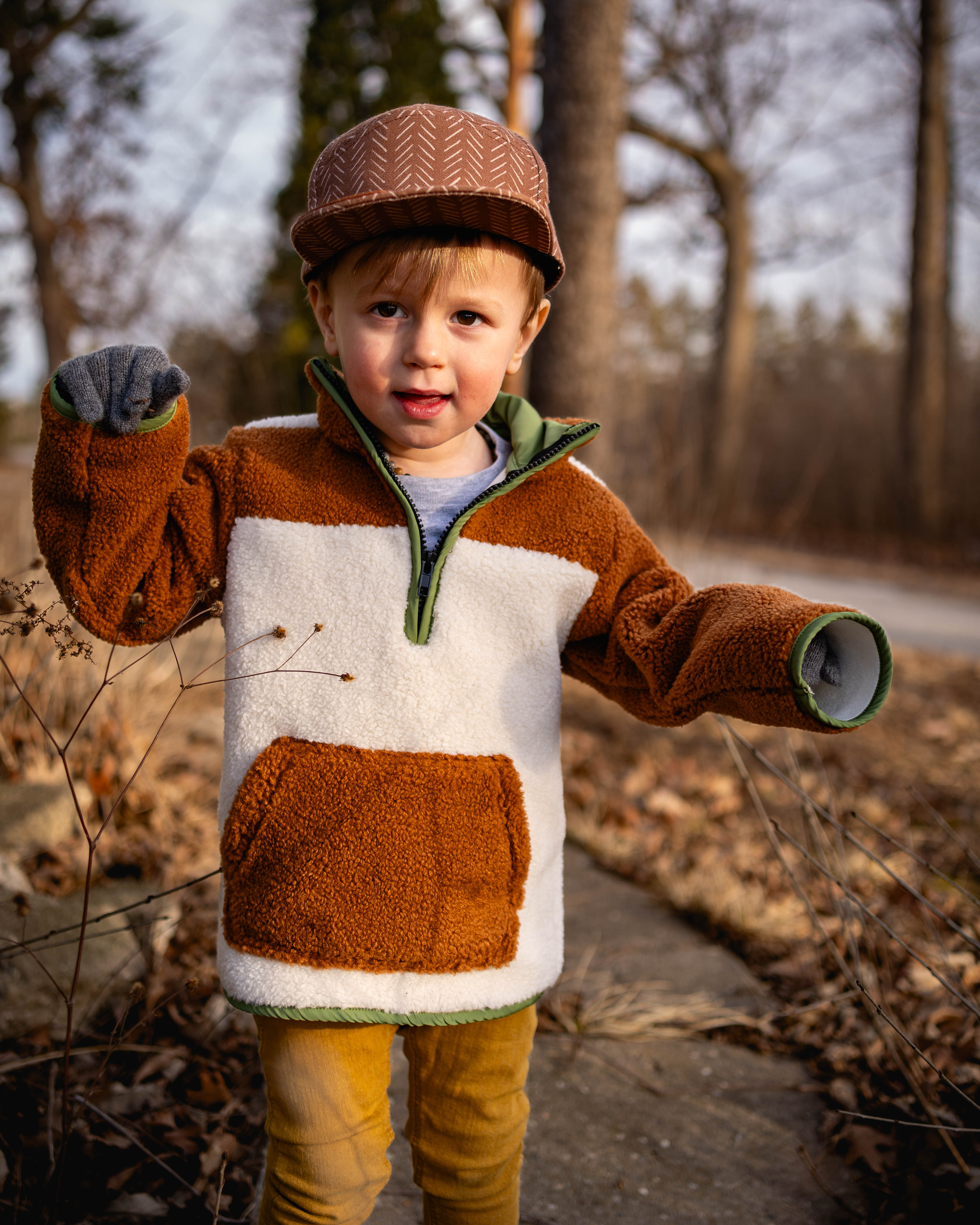Child wearing a brown and white jacket with a knit cap outdoors on a path with trees.