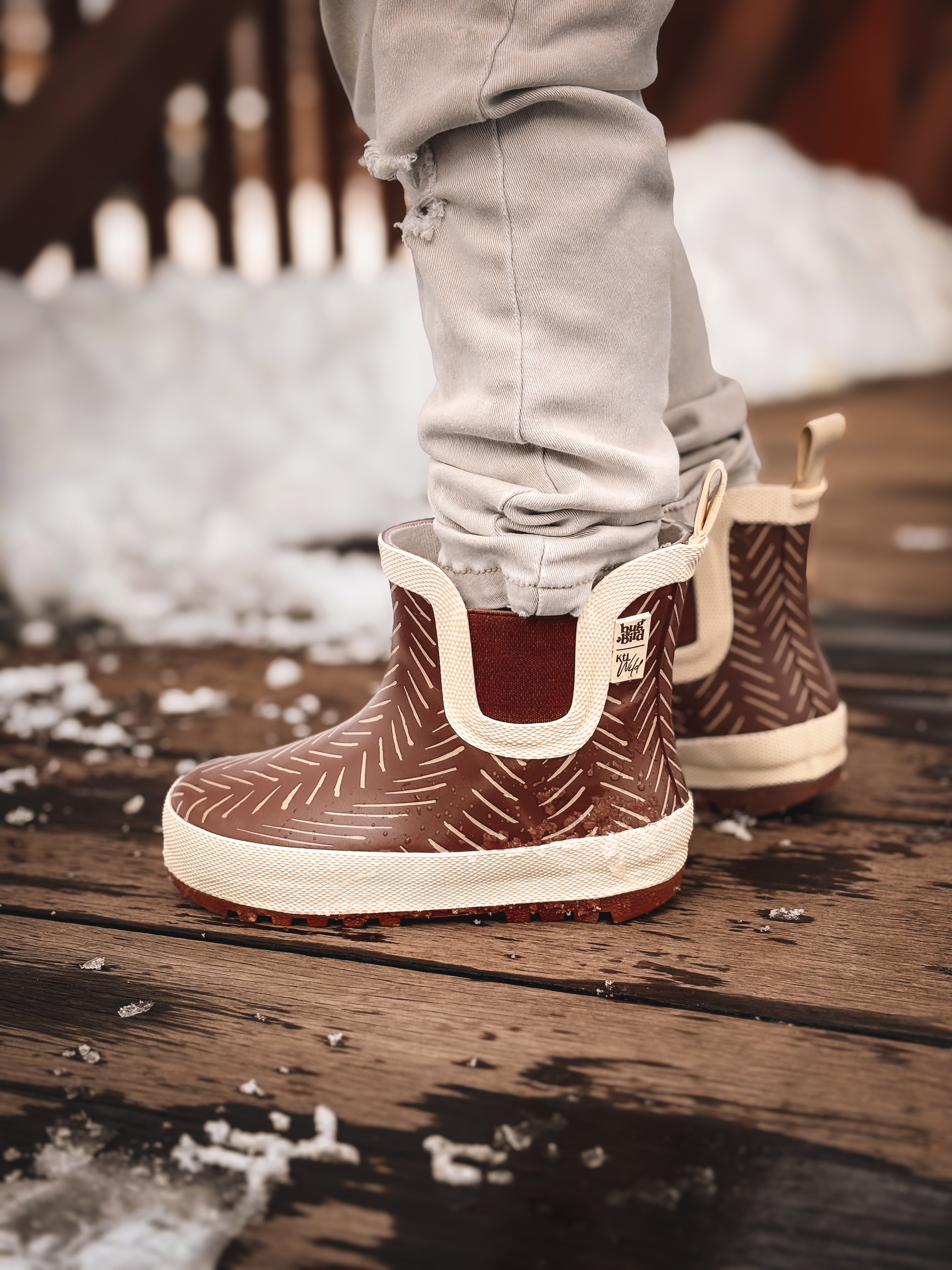 Brown rain boots with white soles worn by a person on a wooden deck.