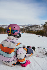 Person in colorful snowboarding gear sitting on a snowy slope with a mountainous background