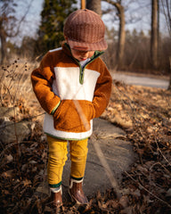 Child wearing a brown and white jacket, yellow pants, and a brown hat standing on a path in a forest.