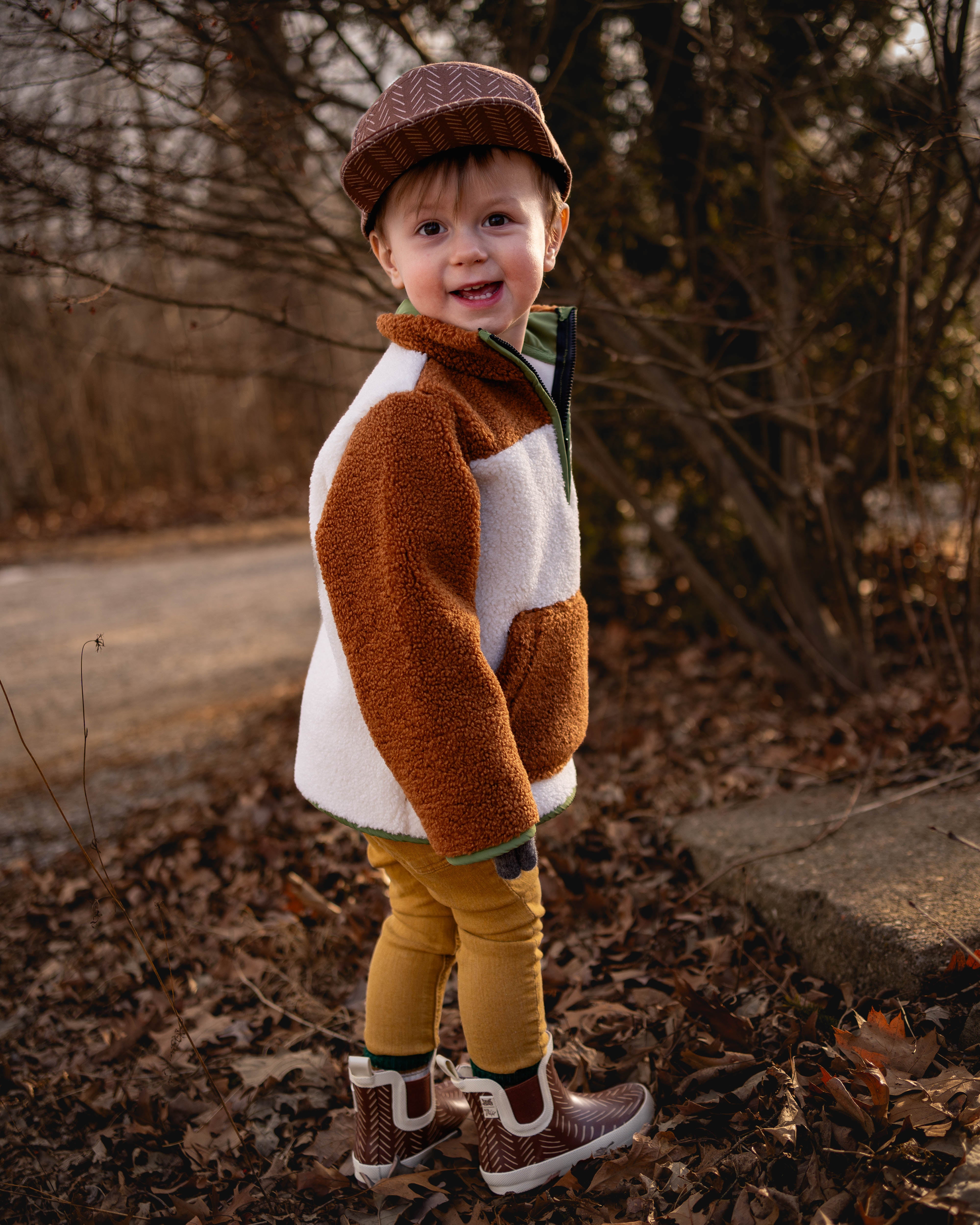 Child wearing a brown and white jacket, yellow pants, and a hat standing outdoors with bare trees in the background.