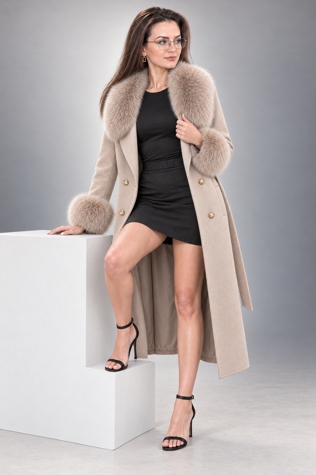A woman models a long beige coat with a large beige fur collar and cuffs, styled with a black mini dress and black high-heeled sandals, standing on a white pedestal against a gray studio background.