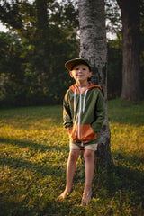 Child wearing a green and orange hoodie standing next to a tree in a grassy area