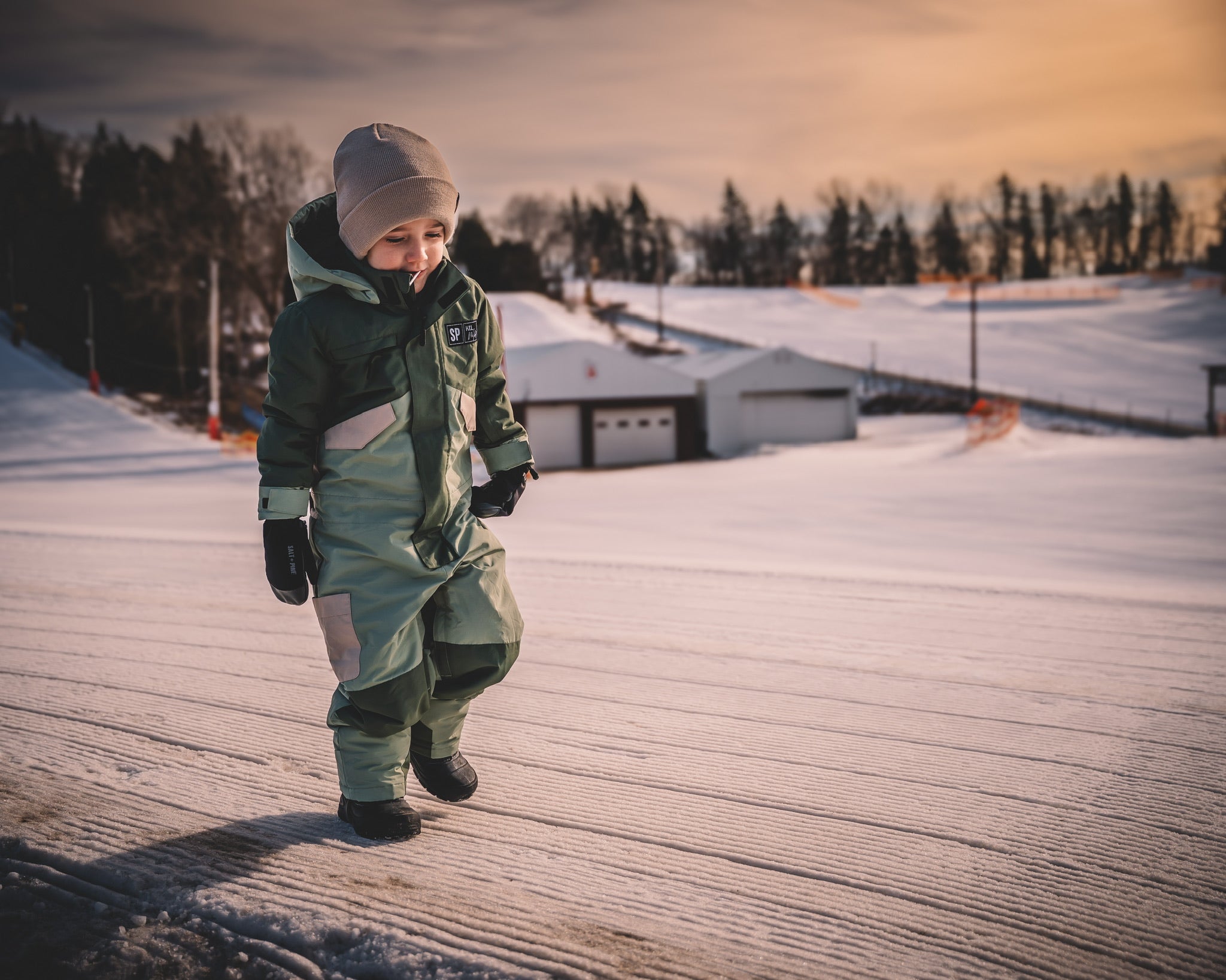 Child in winter clothing walking on a snow-covered path with a snowy landscape and trees in the background.