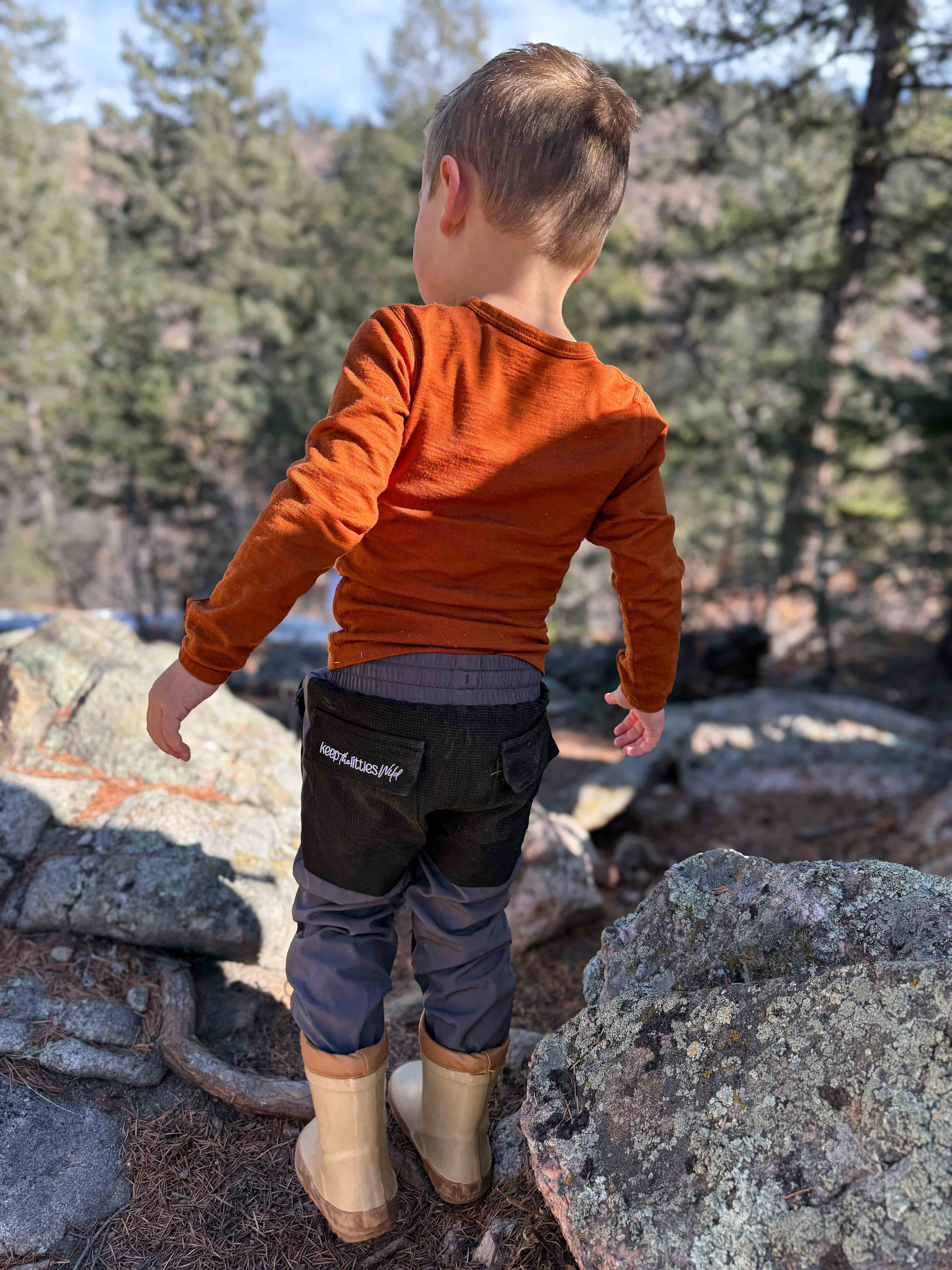 Child in orange shirt and black pants standing on rocks with trees in the background
