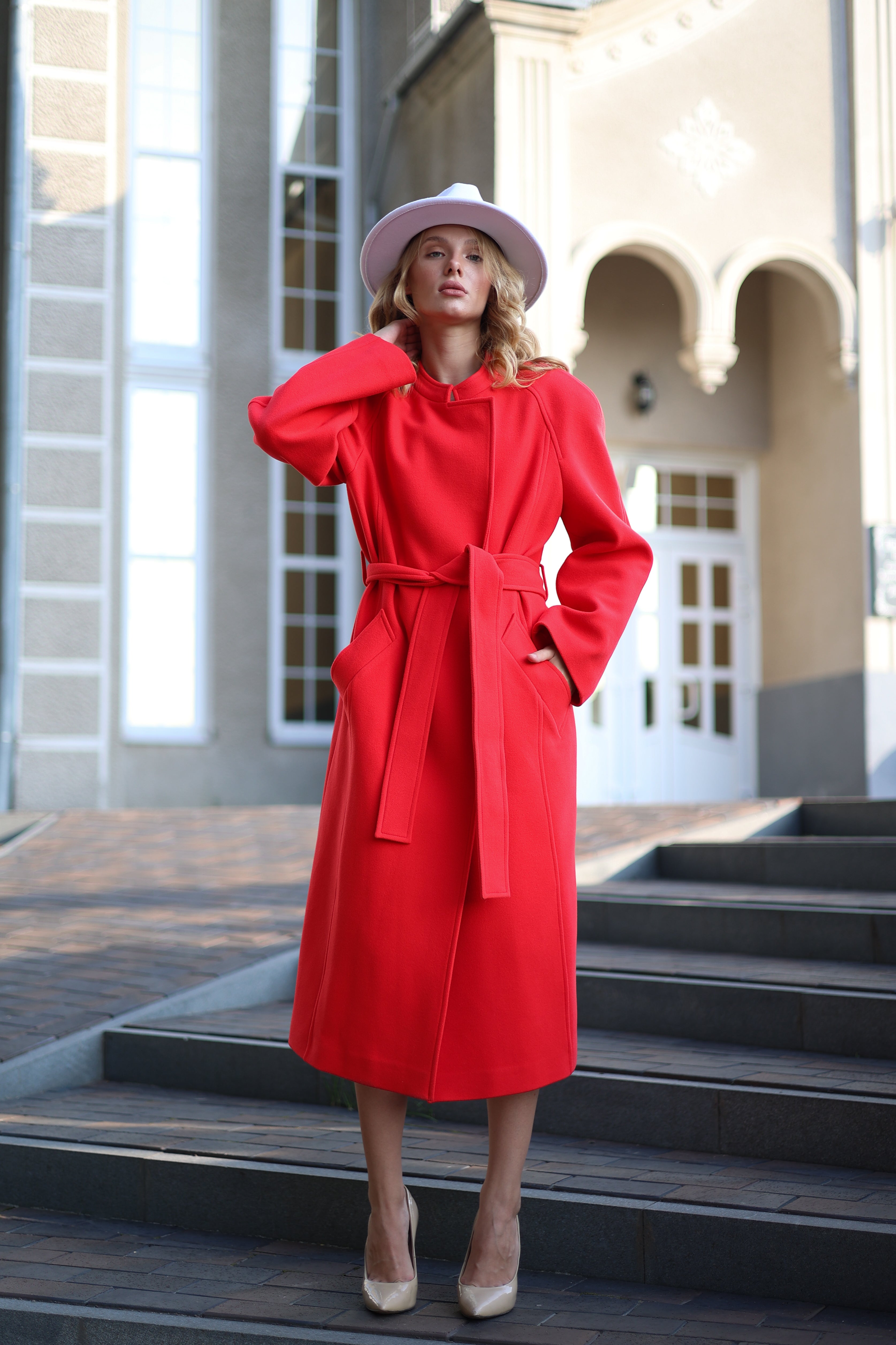 Woman wearing a red coat and white hat standing on steps in front of a building.