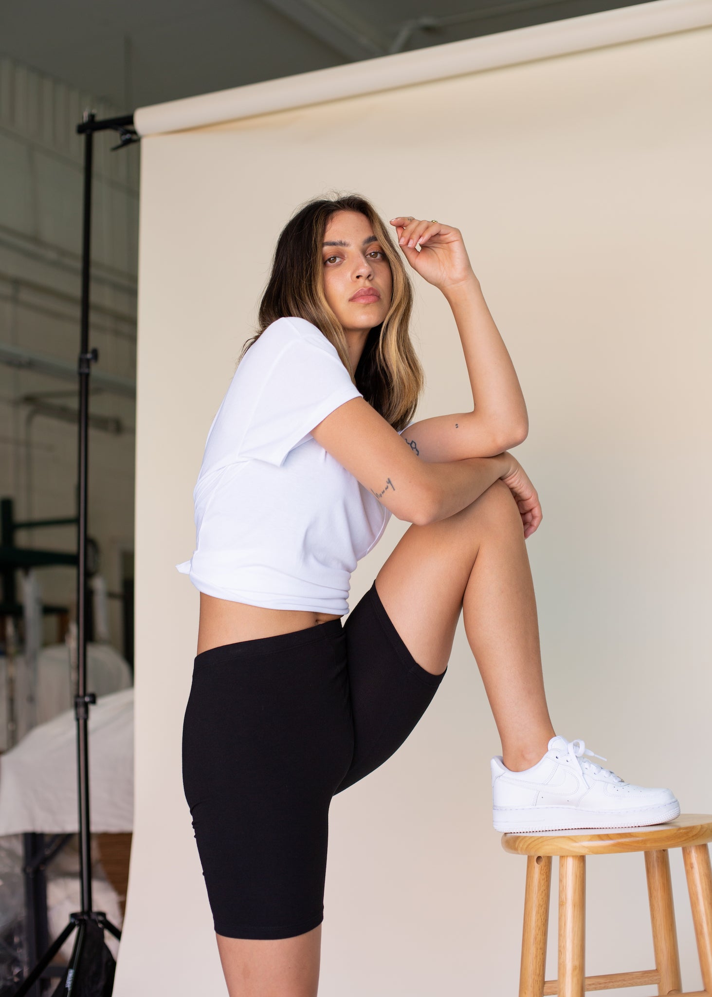Woman in a white crop top and black shorts sitting on a stool indoors.