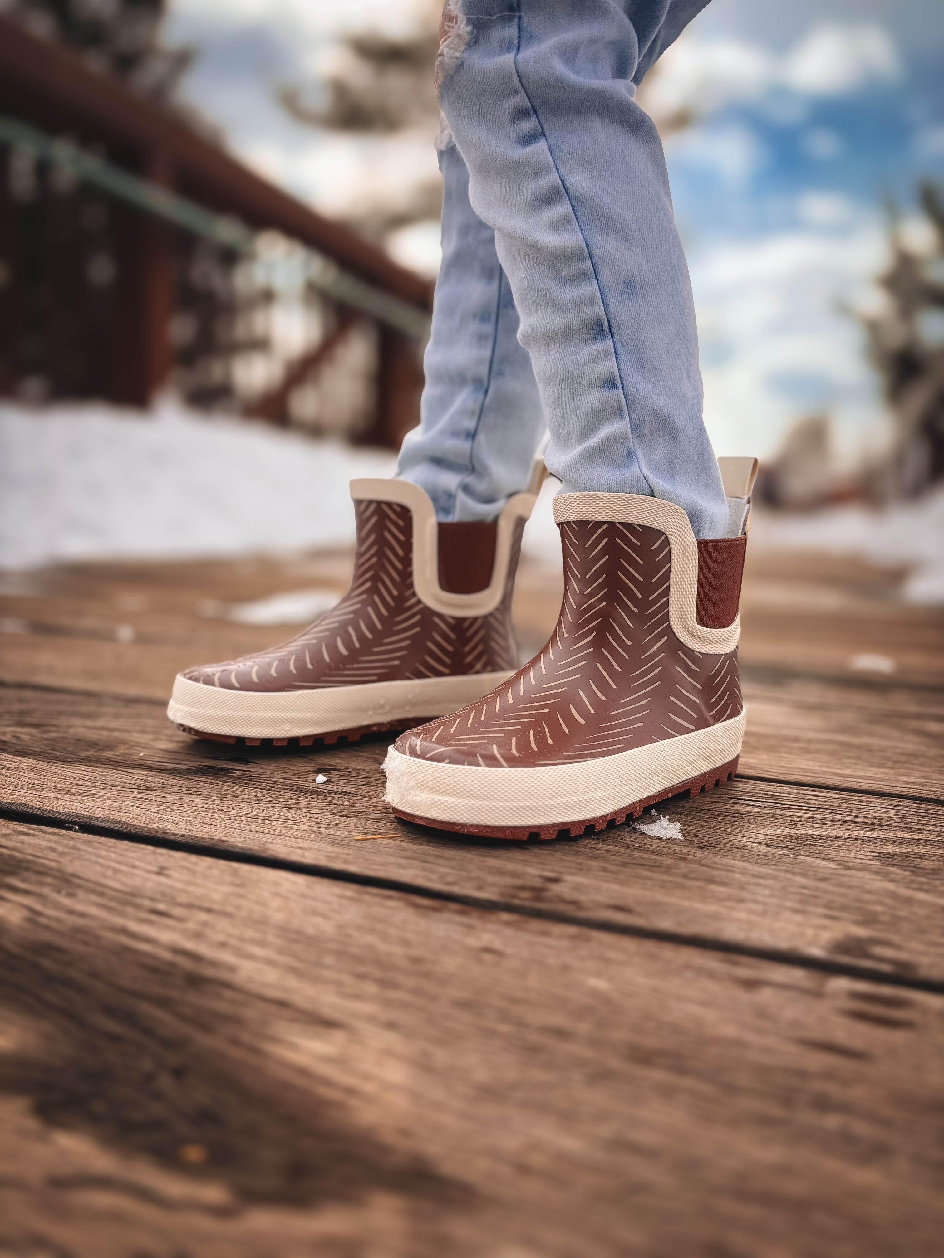 Brown and white patterned rain boots on a wooden floor with a blurred outdoor background.