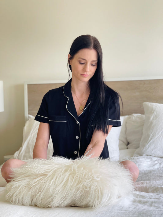 Woman in black pajamas sitting on a bed with a fluffy white pillow.