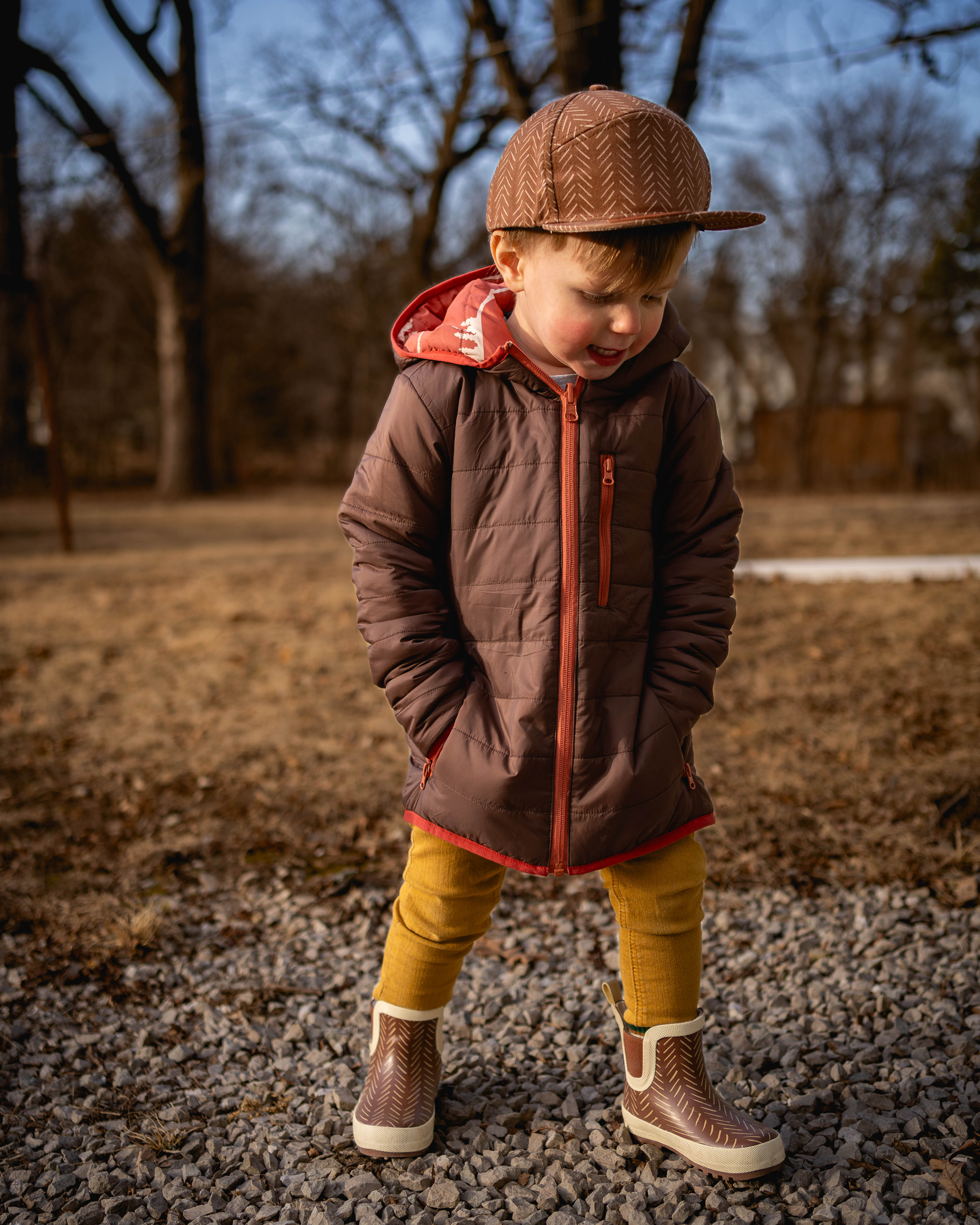 Child wearing a brown jacket, yellow pants, and a cap outdoors on a gravel path with trees in the background.