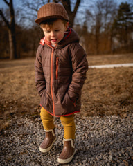 Child wearing a brown coat and hat standing on a gravel path with trees in the background
