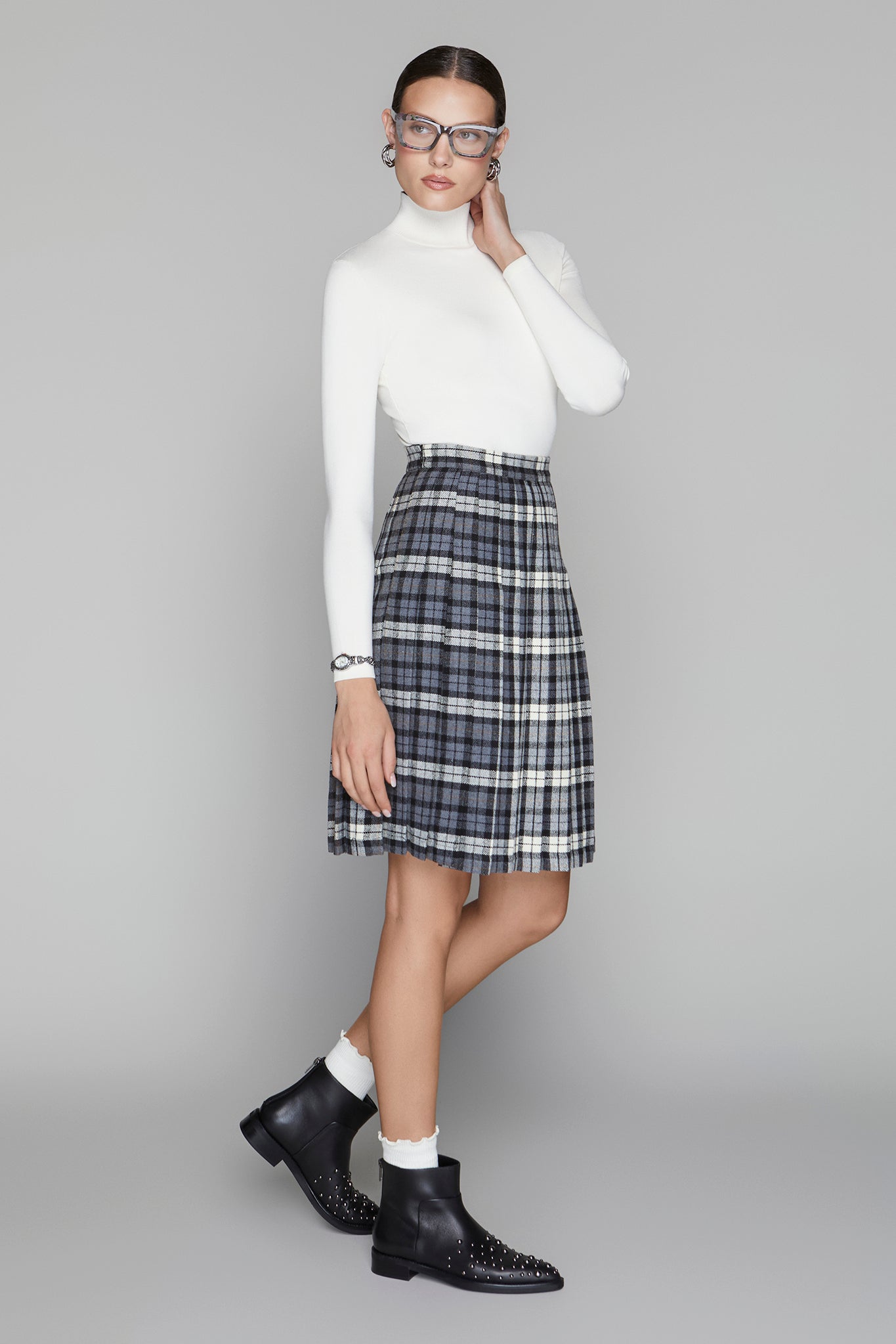 Woman wearing a white turtleneck, plaid skirt, and black boots on a gray background