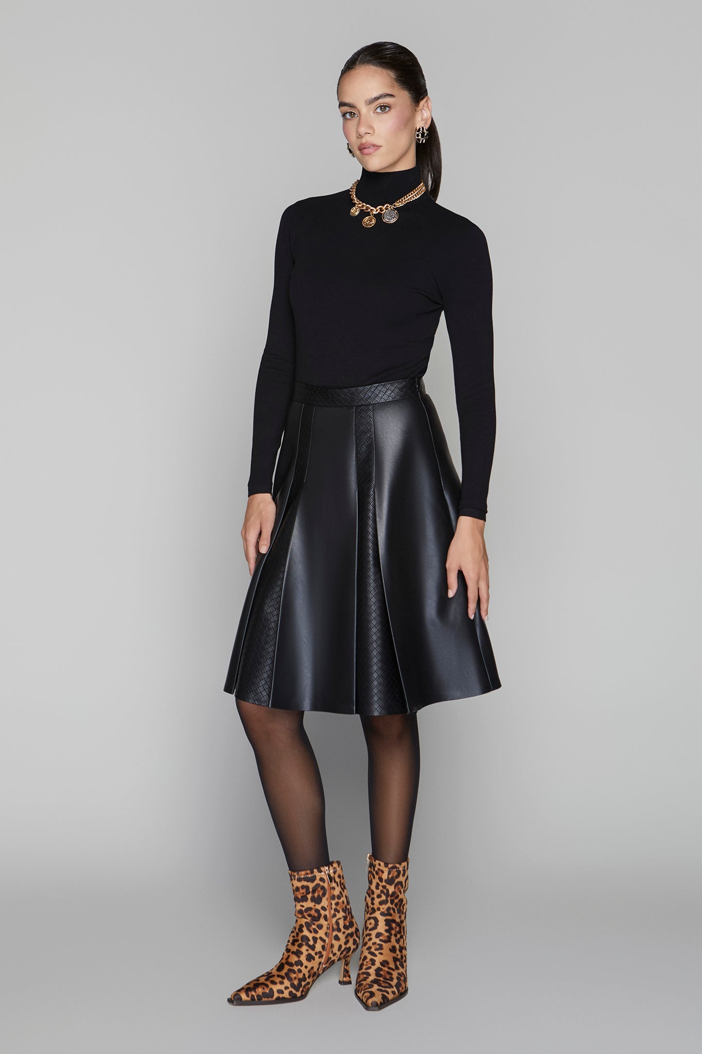 Woman wearing a black turtleneck, black leather skirt, and leopard print boots on a gray background