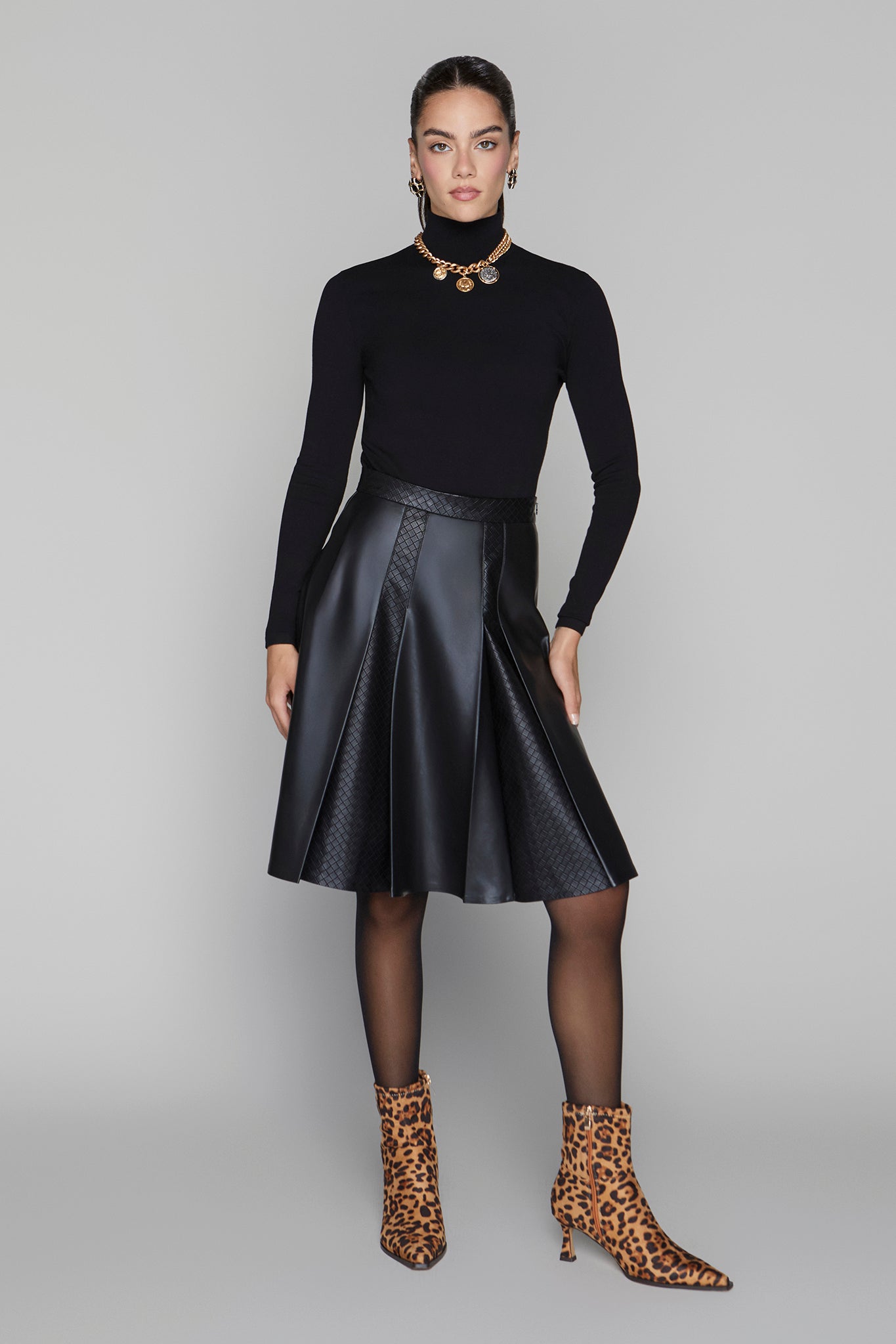 Woman wearing a black turtleneck, black leather skirt, and leopard print boots on a gray background
