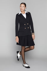 Woman wearing a black blazer with gold buttons and a black pleated skirt on a gray background