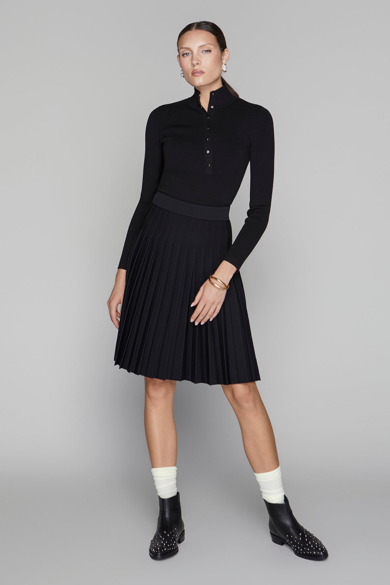 Woman wearing a black long-sleeve top and skirt with white socks and black shoes on a gray background