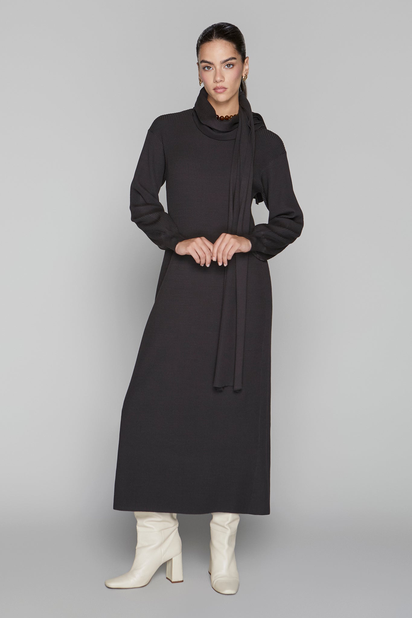 Woman wearing a long black dress with a cowl neck and white boots on a gray background