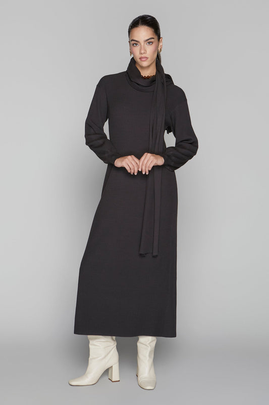 Woman wearing a long black dress with a cowl neck and white boots on a gray background