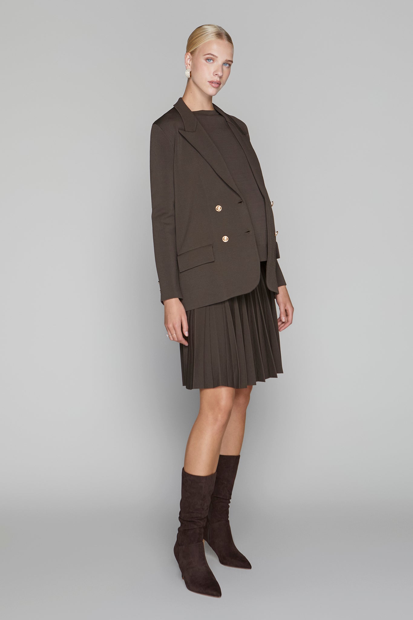 Woman wearing a brown blazer, pleated skirt, and knee-high boots on a gray background