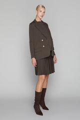 Woman wearing a brown blazer, pleated skirt, and knee-high boots on a gray background