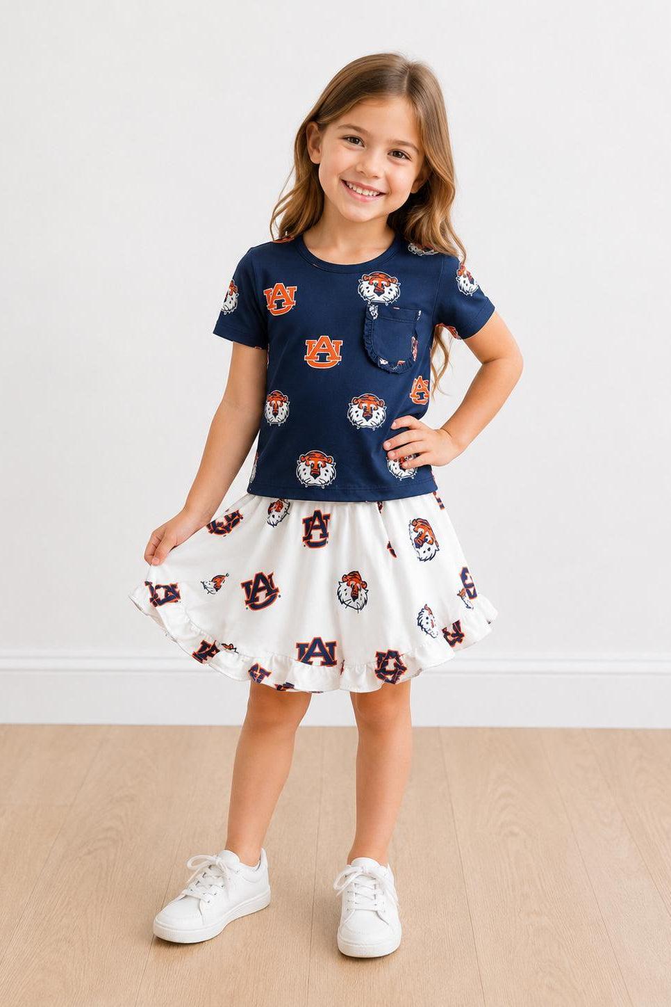 Young girl wearing a navy blue top with a pocket and a white skirt with a pattern, standing on a beige floor.