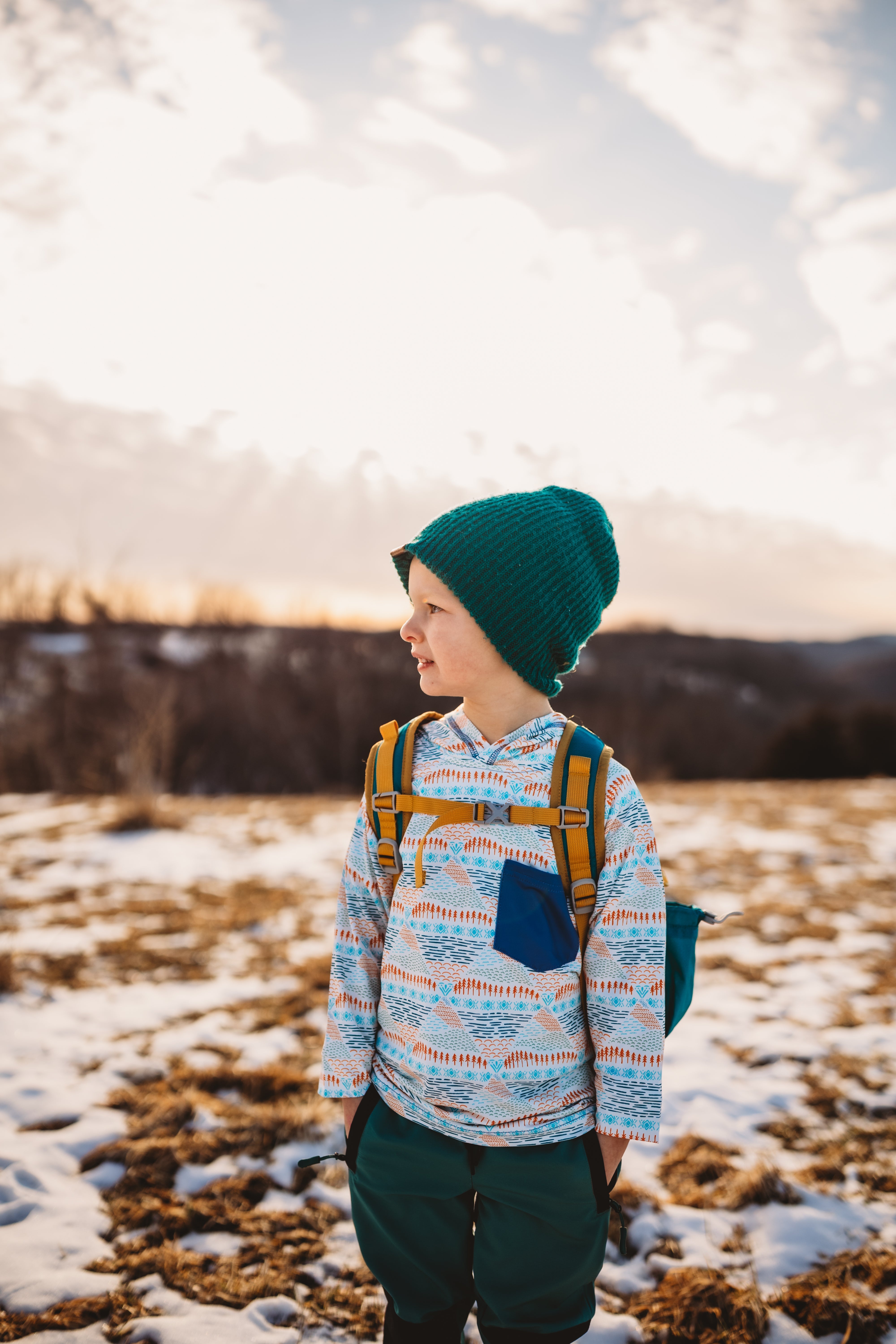 Person wearing a colorful jacket and green beanie standing in a snowy landscape with mountains in the background.