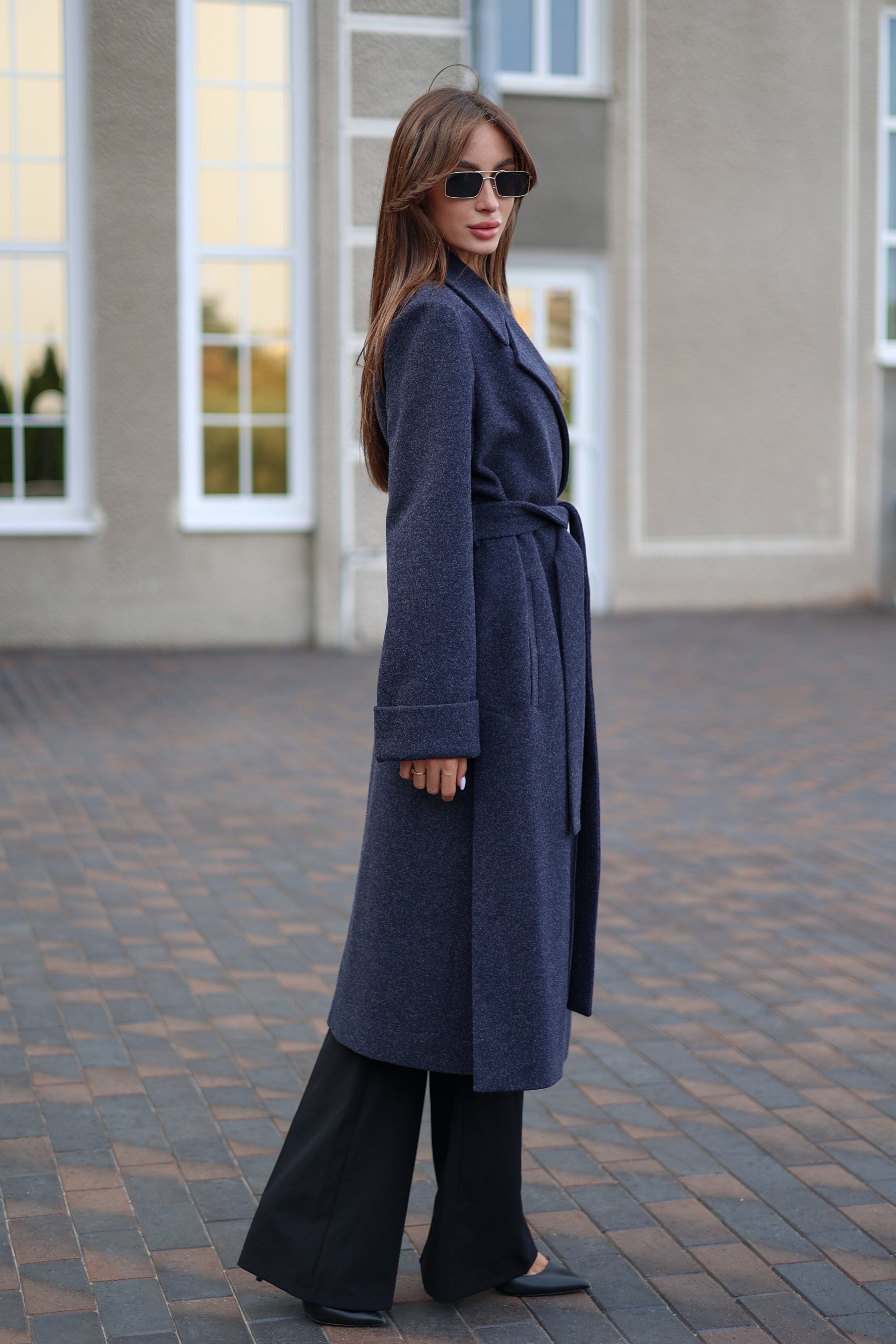 Woman wearing a long blue coat standing on a paved area with a building in the background