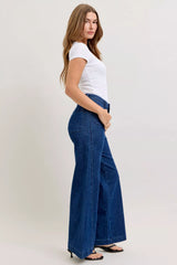 Woman wearing blue wide-leg jeans and a white top on a plain background