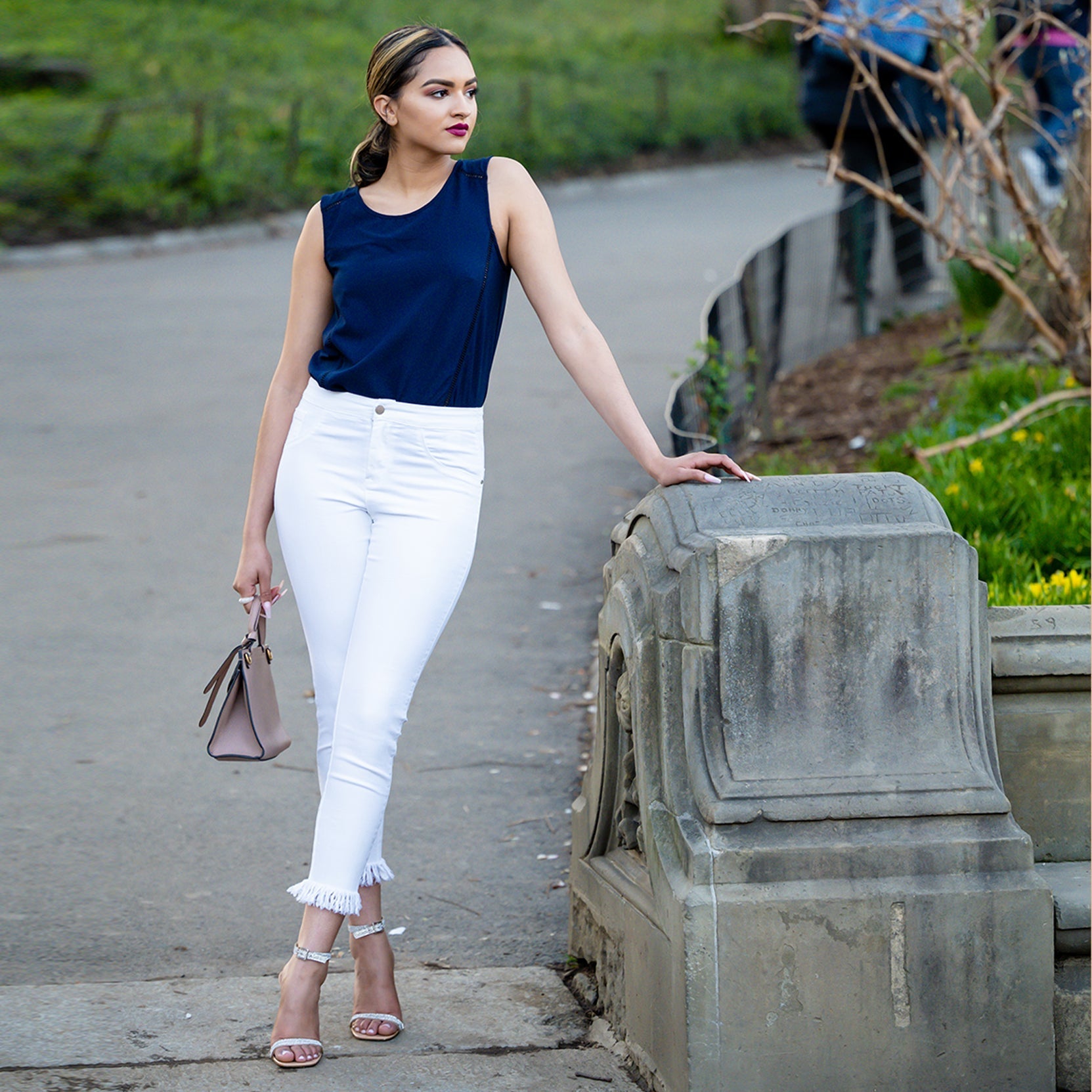 Woman in navy top and white pants standing on a path with greenery in the background